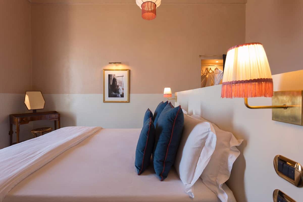 A hotel room with a bed, bedside lamps, a framed picture on the wall, and a small table with a lamp.