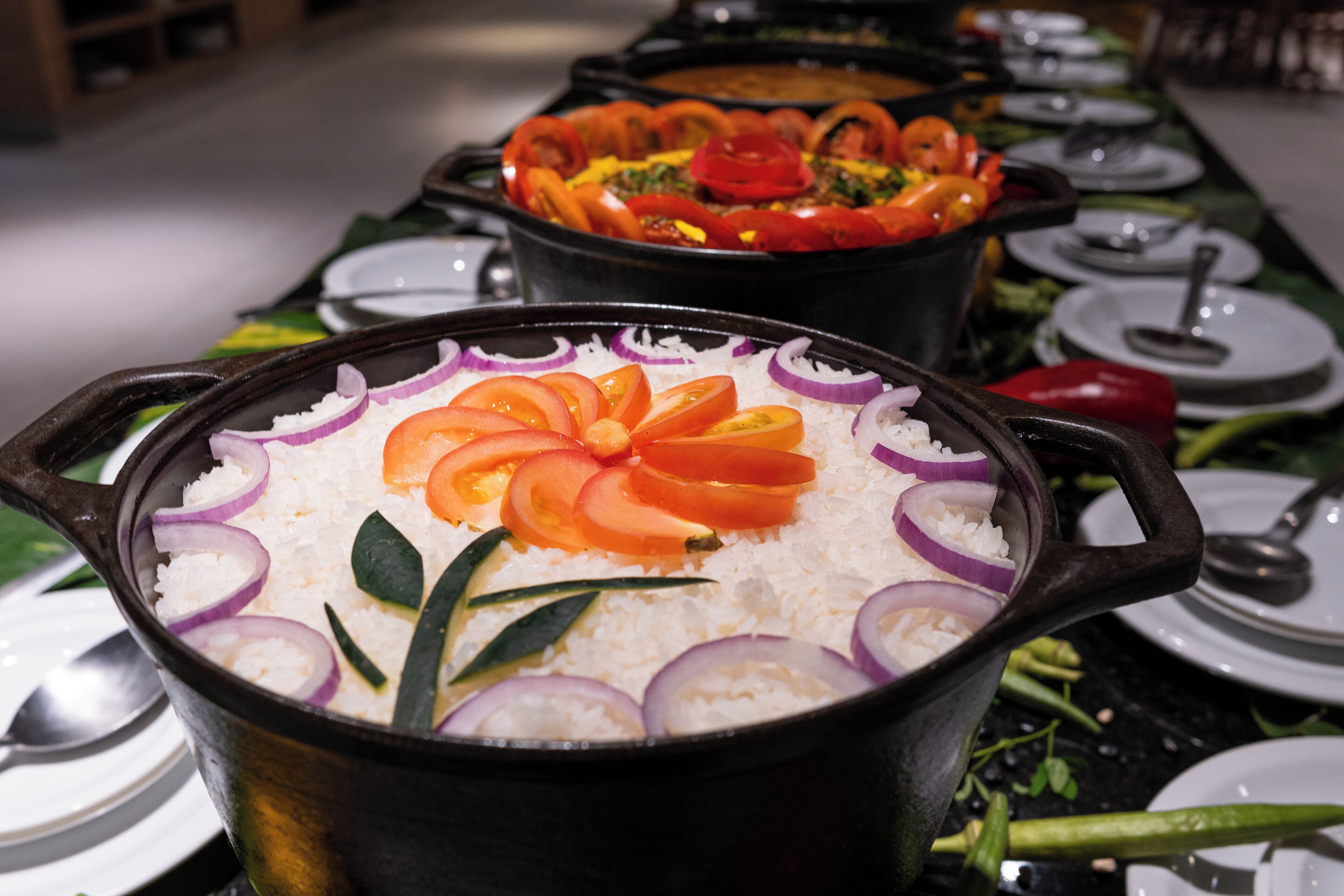 Uma mesa de buffet com diversos pratos, incluindo uma panela de arroz guarnecida com tomate e cebola, e outro prato com legumes e carne.