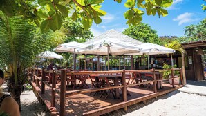 An outdoor dining area with wooden tables and chairs, covered by large umbrellas, and surrounded by lush greenery. - Ondas Praia Resort by WAM Experience  (Porto Seguro)