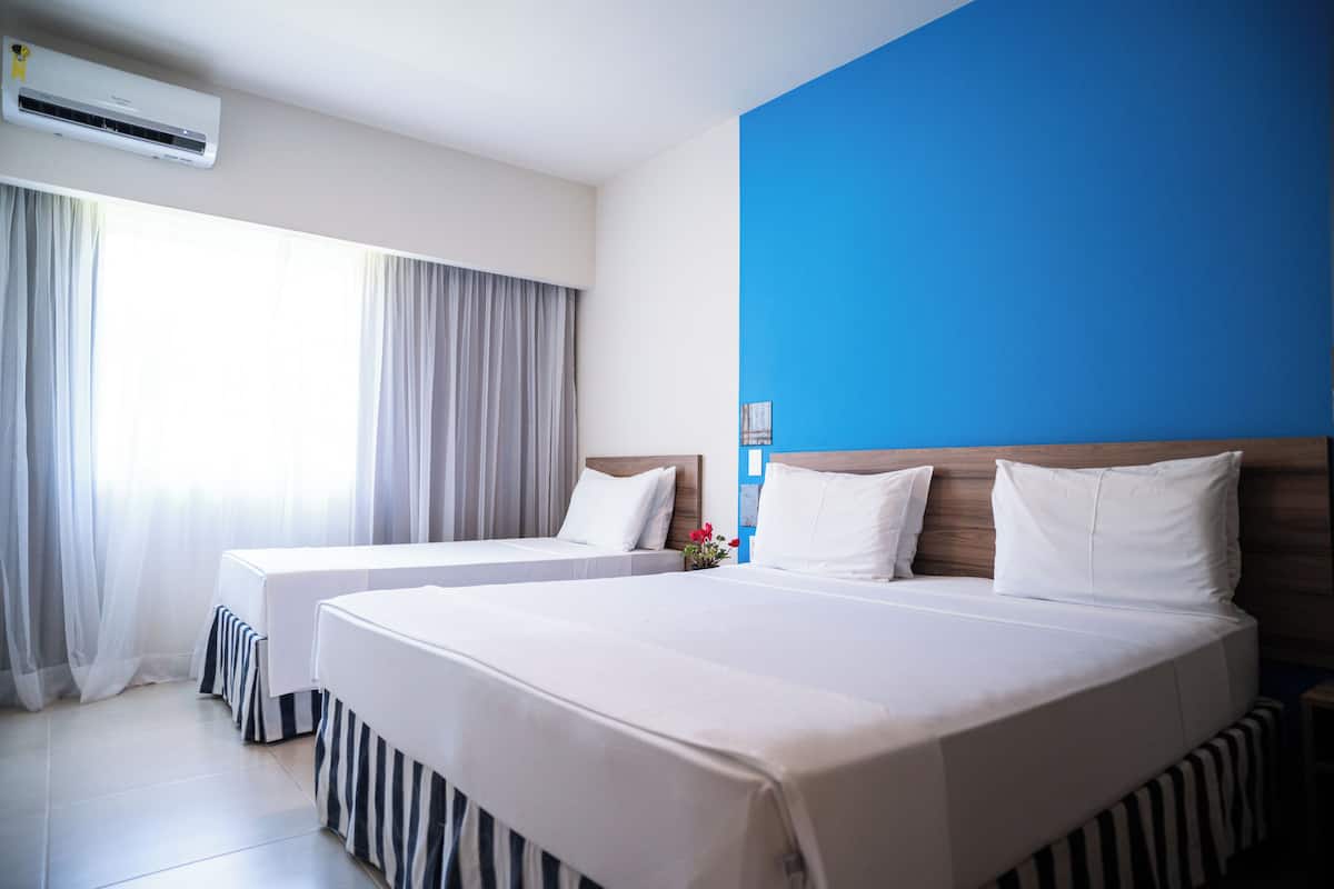 Ein Hotelzimmer mit zwei Betten, einem großen Fenster mit Vorhängen, einer Klimaanlage und einer blauen Akzentwand.