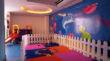 Área de recreação infantil com cerca branca de piquete, tapetes de atividades coloridos e um mural com criaturas marinhas.