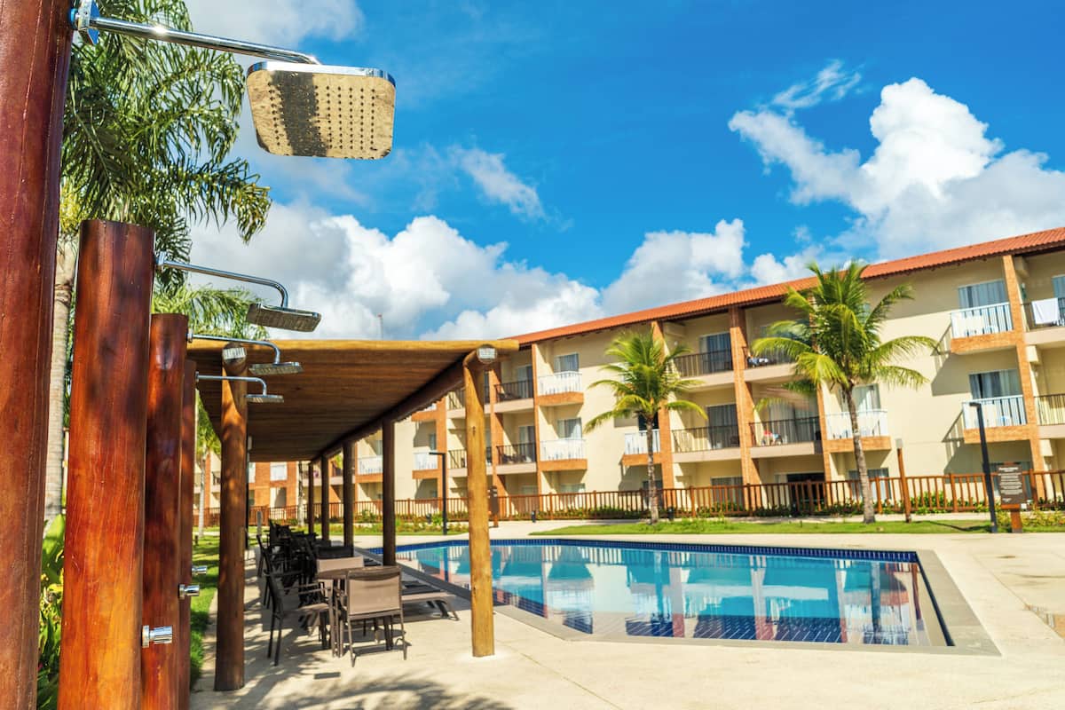 Ein Poolbereich mit überdachter Sitzgelegenheit, einer Holzpergola und einem Gebäude mit Balkonen sowie Palmen im Hintergrund.