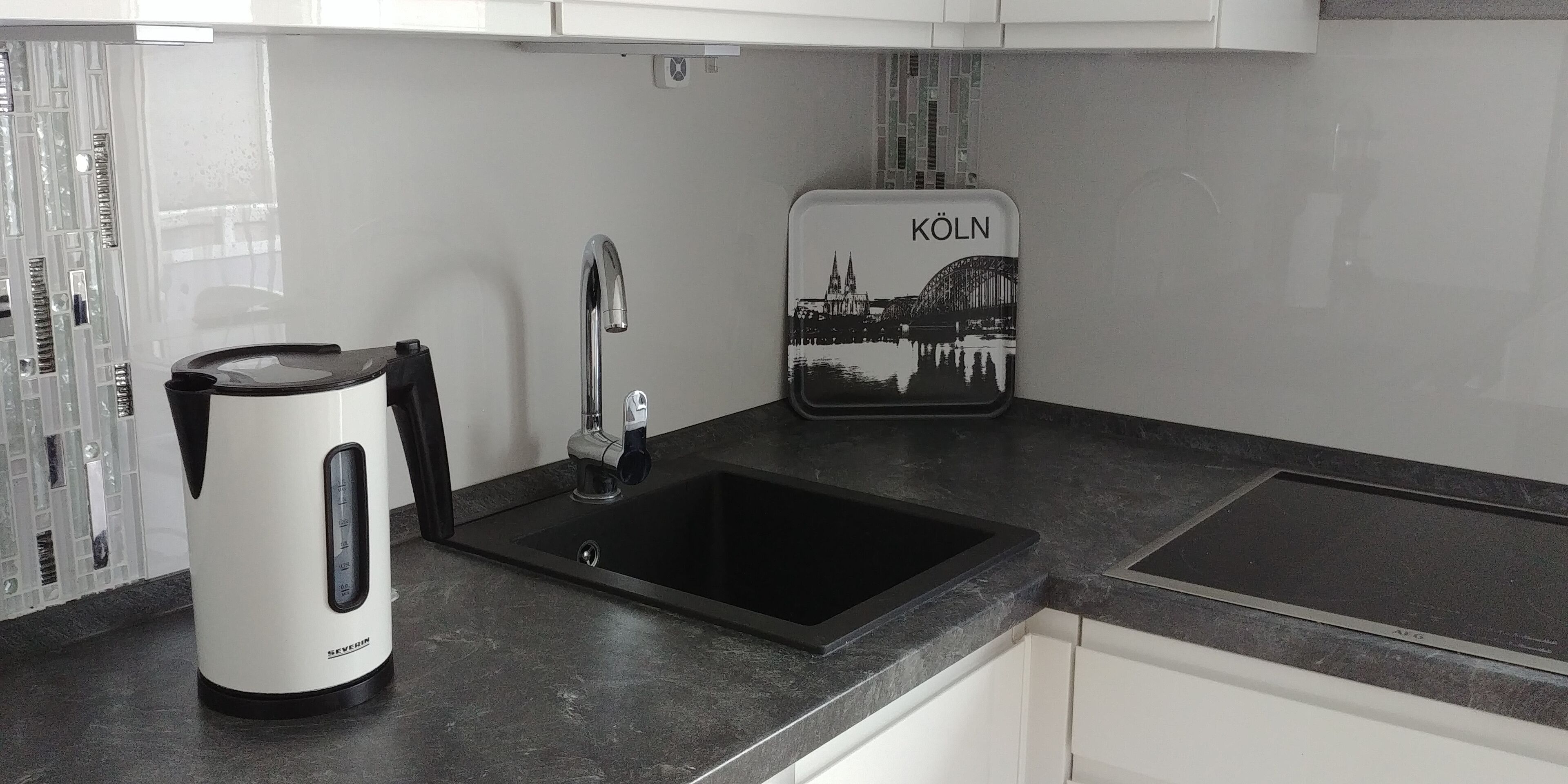 A kitchen with a black countertop, a white kettle, a sink, and a black and white cityscape sign.