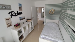 A bedroom with a bed, a white dresser, a shelf with decorative items, and a wall with a 'Relax' sign.