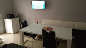A small, modern living room with a white table, black chairs, a white sofa, a flat-screen TV mounted on the wall, and a microwave on a small white cabinet.