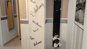 A hallway with a wall-mounted sign listing wellness-related terms, a mirror, a door, a black towel rack, a framed photo, and a small potted plant.