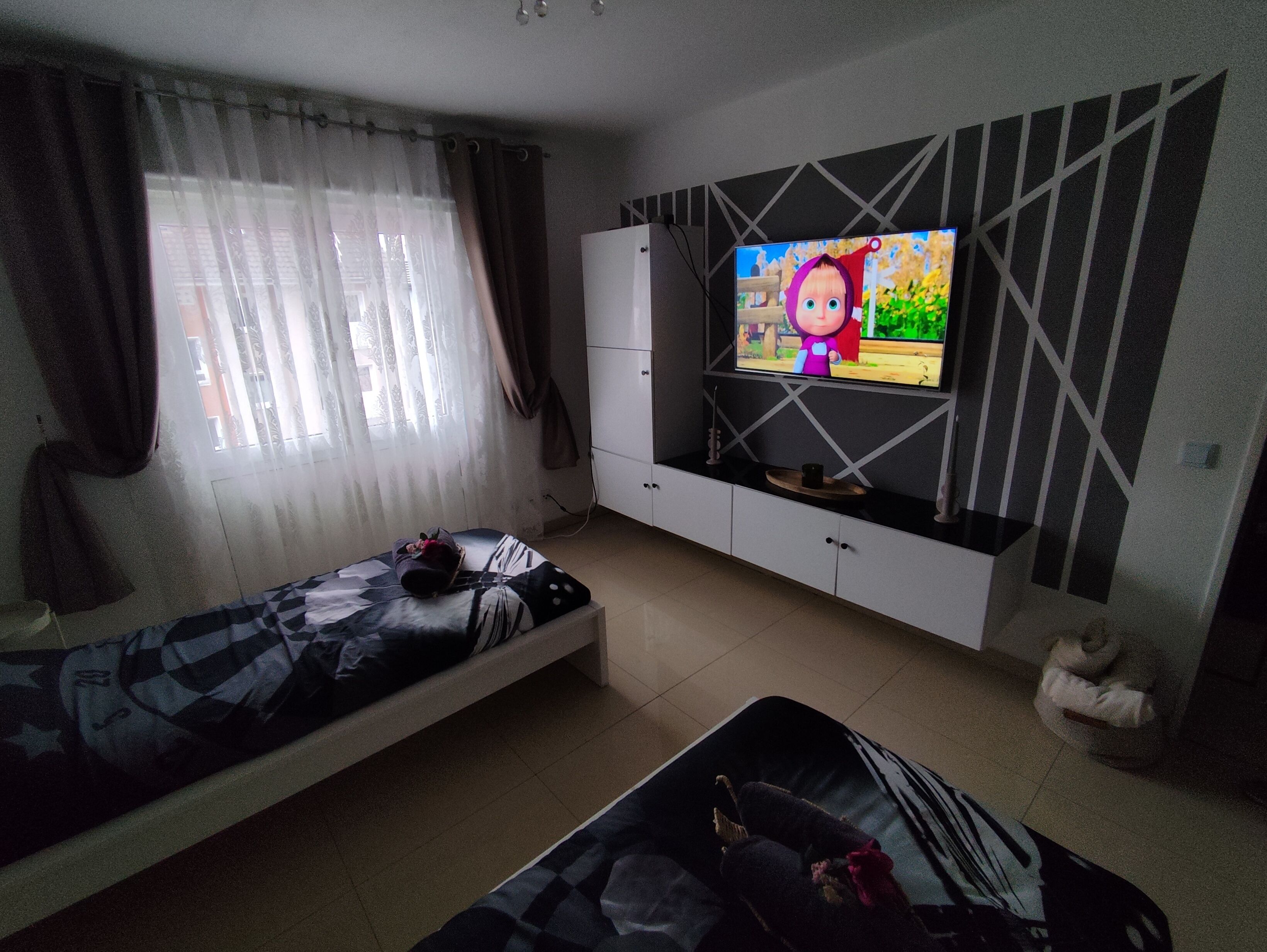A bedroom with a bed, a TV mounted on the wall, a wardrobe, and a window with curtains.