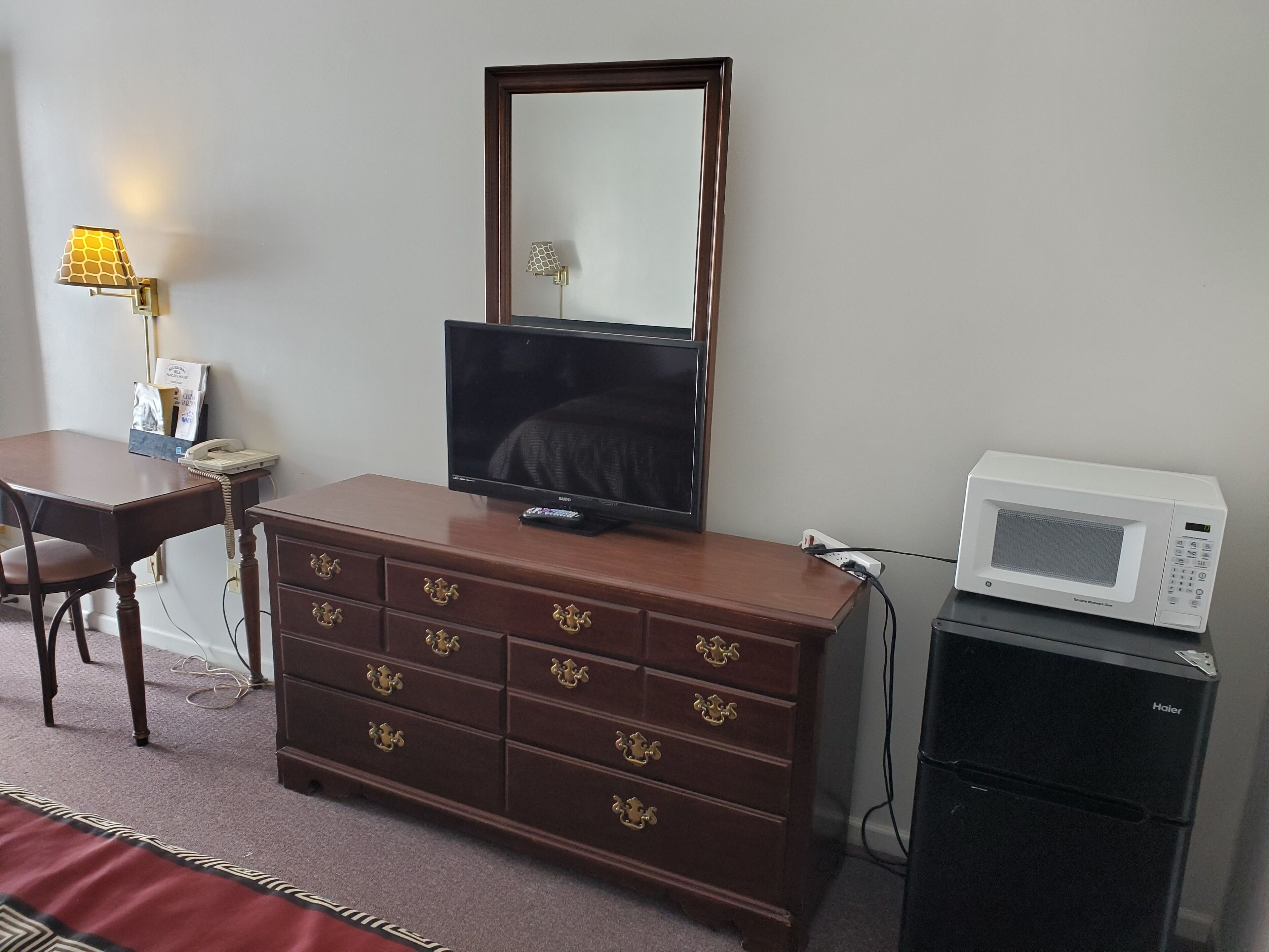 A hotel room with a wooden dresser, a television, a mirror, a lamp, a telephone, and a microwave.