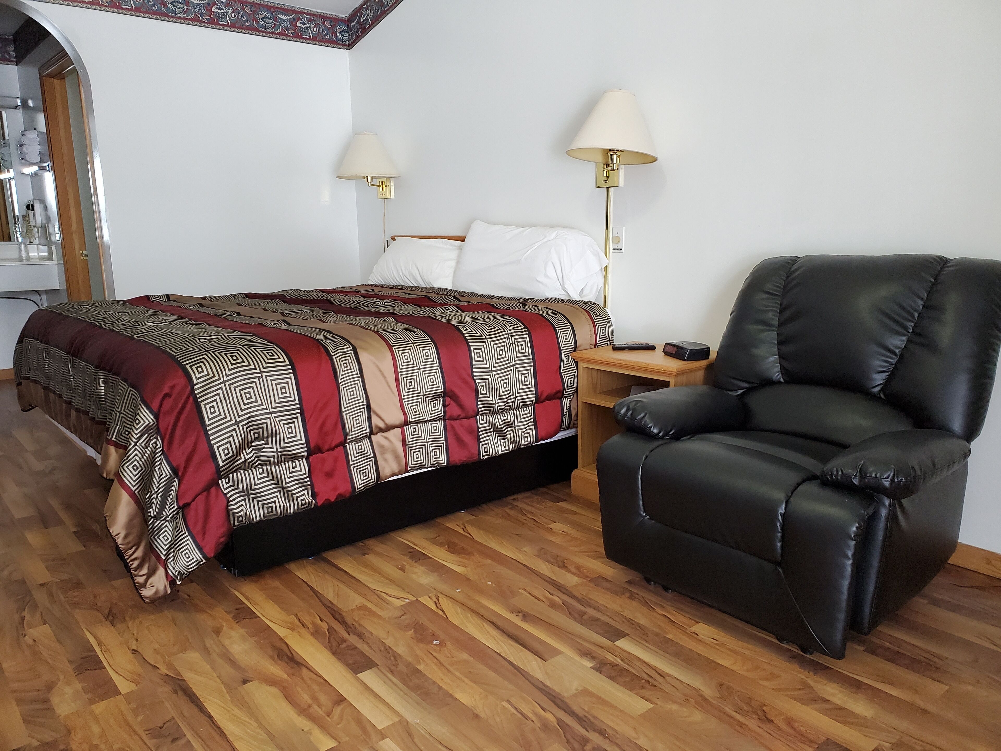 Un dormitorio con suelo de madera, una cama con colcha estampada, dos lámparas de noche, un sillón de cuero negro y una mesita de noche de madera.