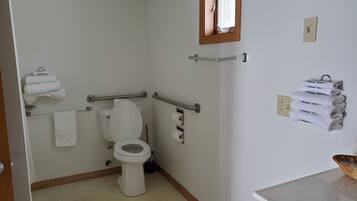 A small, clean bathroom with a toilet, handrails, a towel rack, and a window.