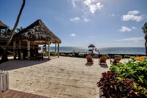 A beachside patio with thatched-roof structures, wooden chairs, and a view of the ocean.