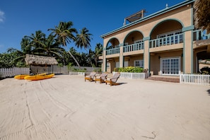 A beachfront property with a large building, a thatched-roof structure, and a fenced area with lounge chairs and a yellow kayak.