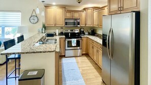A modern kitchen with granite countertops, stainless steel appliances, wooden cabinets, and a dining area with bar stools. - Sunset Beach Getaway – Steps to the Beach, Walk to Restaurants, Sleeps 8! (Treasure Island)