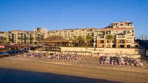 Un hôtel à étages, une plage aménagée avec des parasols et des chaises longues, et une structure surmontée d’un toit de chaume.