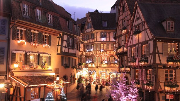 Uma rua europeia tradicional com edifícios de enxaimel, um mercado de Natal e uma árvore de Natal decorada.