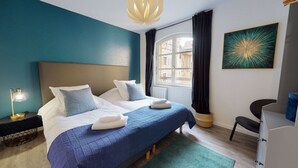 Quarto com duas camas, uma escrivaninha, uma cadeira, uma janela com cortinas e um quadro na parede.