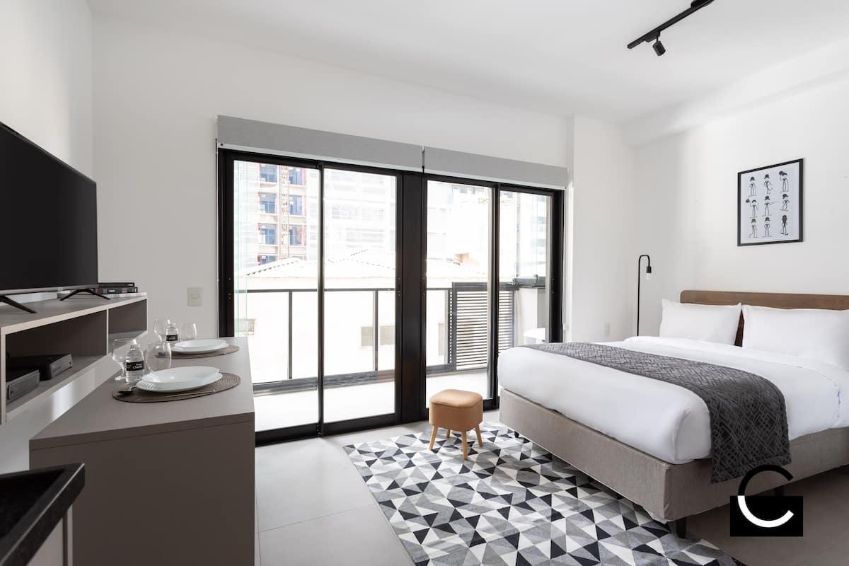Ein modernes Hotelzimmer mit einem Bett, einem Schreibtisch mit Flachbildfernseher, einem kleinen Hocker und einem großen Fenster mit Blick auf die Stadt.