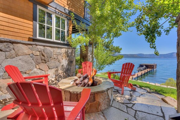 A lakeside patio with red Adirondack chairs, a stone fire pit, and a dock extending into the water.