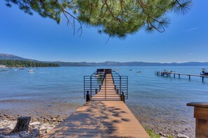 A wooden dock leading to a clear lake with boats, a pier, and mountains in the background.