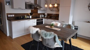 Una cocina moderna con armarios blancos, encimera de madera y una mesa de comedor con sillas. Hay lámparas colgantes sobre la mesa.