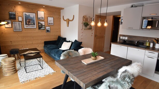 Una sala de estar acogedora con un sofá de madera, una mesa de comedor con una planta y una cocina con armarios blancos y un microondas.