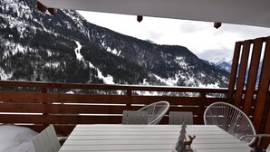 Una terraza de madera con una mesa y dos sillas blancas, con vistas a un paisaje montañoso nevado.