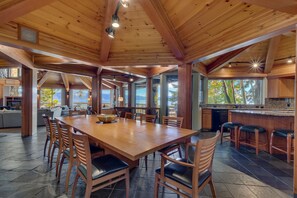 A spacious dining area with a large wooden table and chairs, a kitchen with a bar, and large windows offering views of the outdoors.