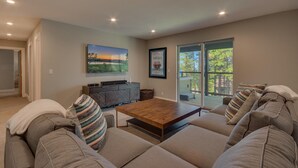 A spacious living room with a large sofa set, a wooden coffee table, a flat-screen TV mounted on the wall, and a sliding glass door leading to a deck.