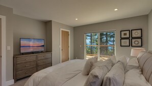 A bedroom with a large bed, a TV on a dresser, and a balcony with a view of trees.