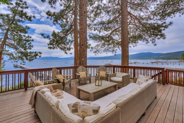 A deck with outdoor furniture, including a sofa, chairs, and a table, overlooking a lake with boats and mountains in the background.