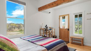 Une chambre avec une grande fenêtre, un lit recouvert d’une couverture aux couleurs vives, un banc en bois et un bureau avec une chaise.