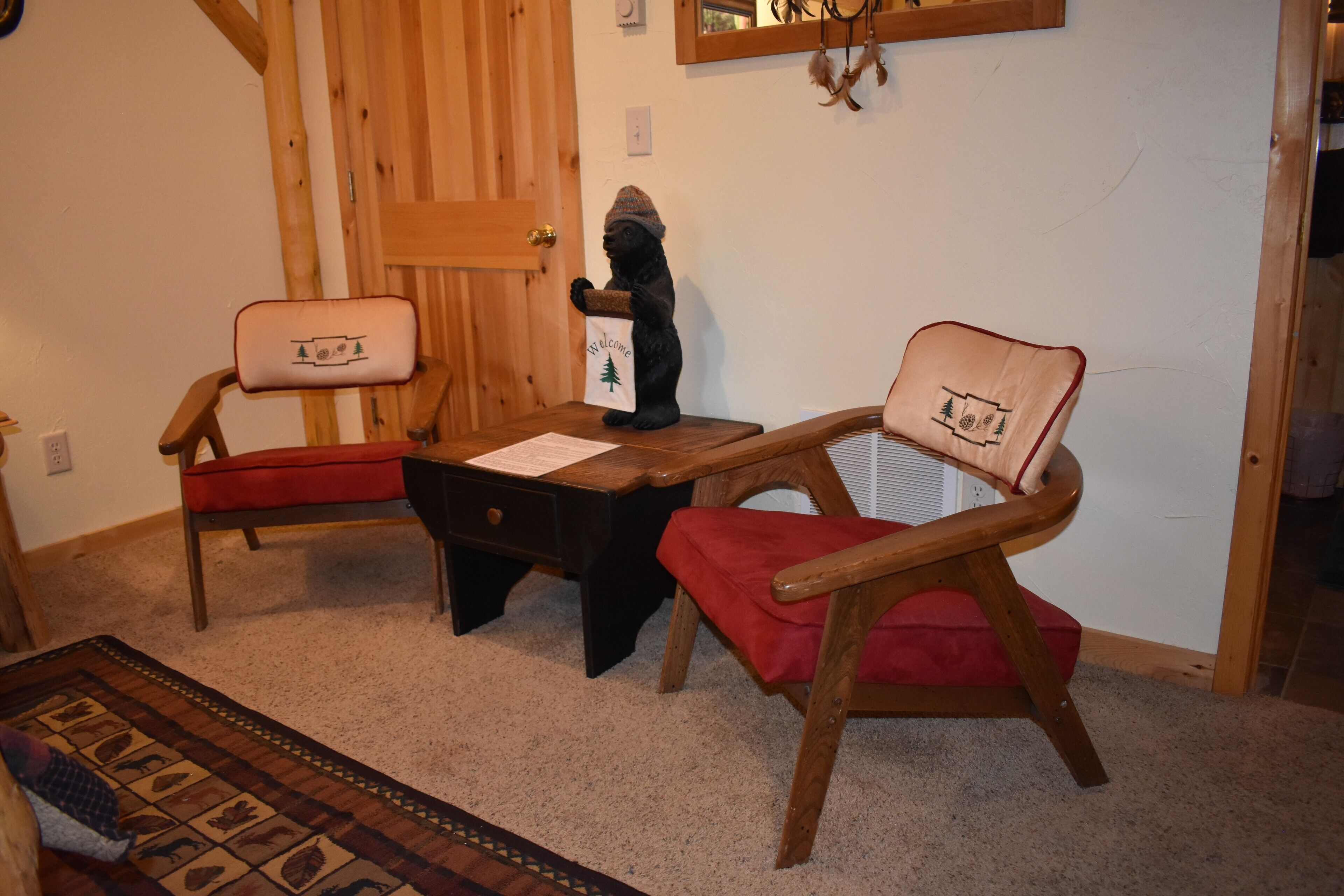 Une pièce comprenant deux chaises en bois avec des coussins rouges, une petite table en bois et une figurine d’ours tenant un sac de bienvenue.
