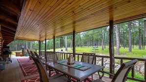 A covered outdoor dining area with a table and chairs, surrounded by a wooden deck and large windows offering a view of a forested area.