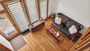 Een moderne woonkamer met een bruine leren bank, houten vloer en grote ramen met zonwering.