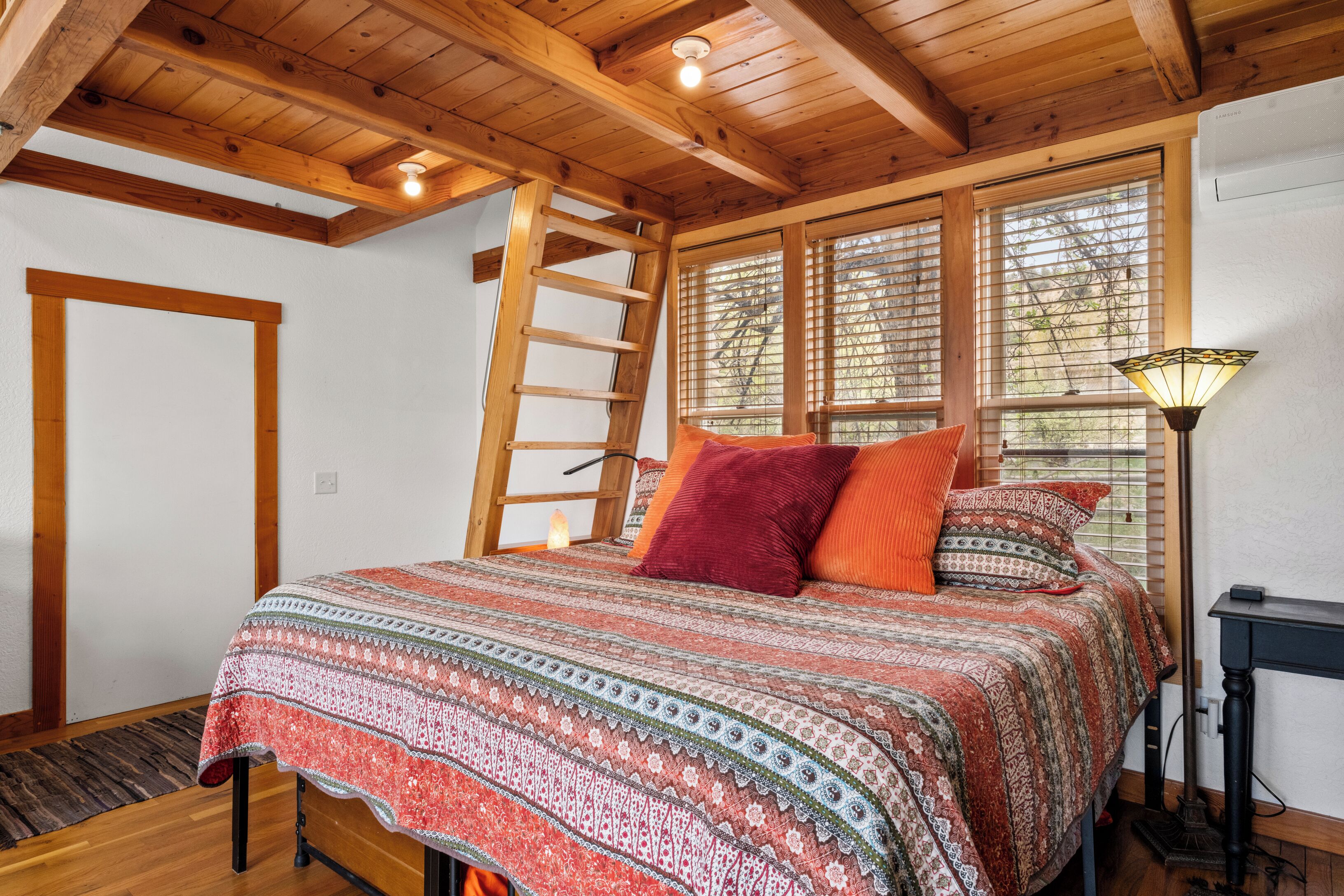 A bedroom with a wooden ceiling, a ladder, a bed with patterned bedding, and a nightstand with a lamp.