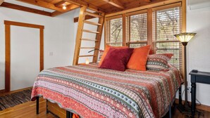 Een slaapkamer met een houten plafond, een ladder, een bed met beddengoed met een patroon en een nachtkastje met een lamp.