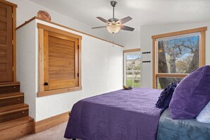Ein Schlafzimmer mit Bett, Holztreppe, Deckenventilator und Fenster mit Blick auf Bäume.