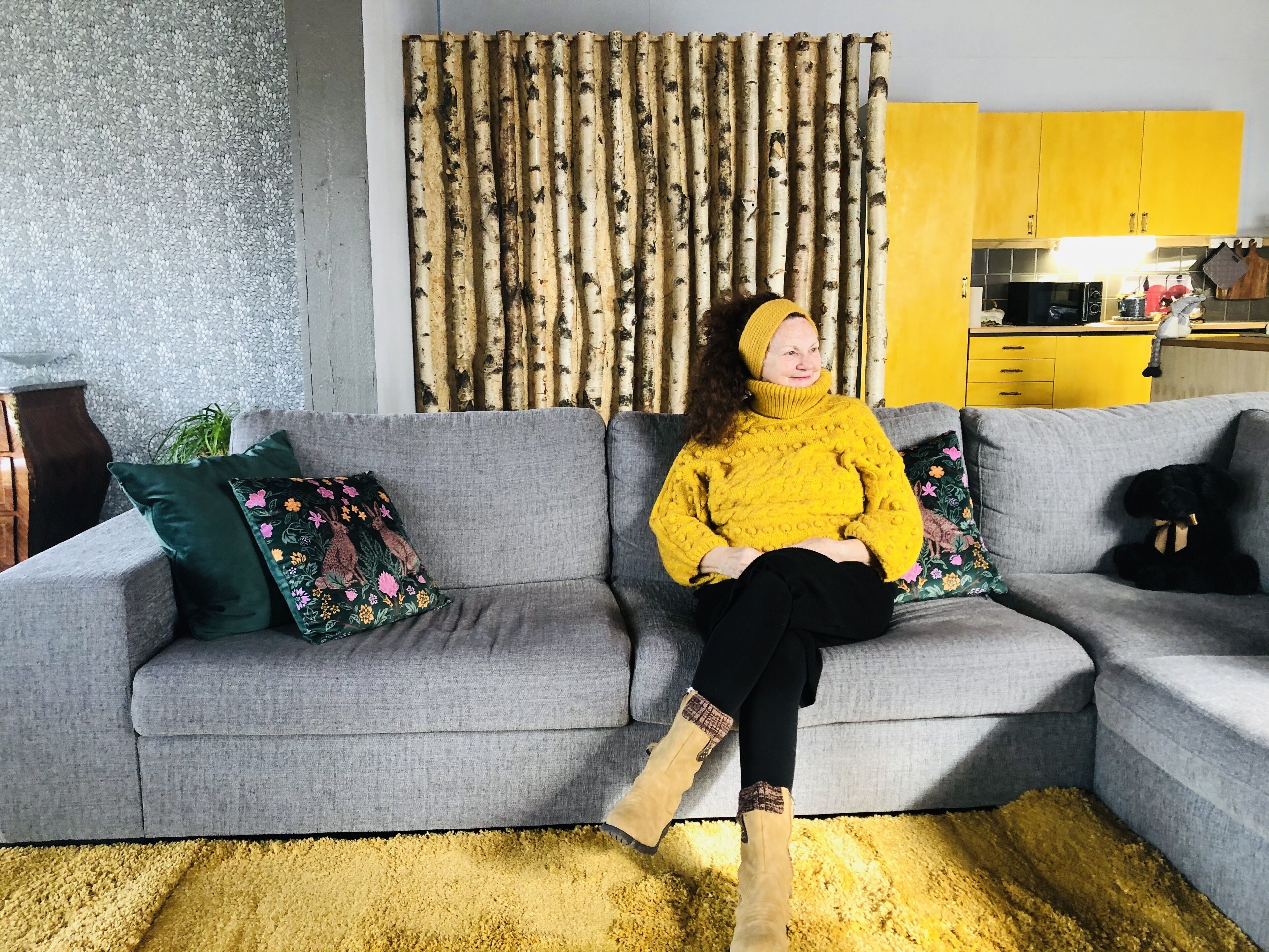 A person sitting on a grey sectional sofa with a yellow rug, a green floral pillow, and a yellow beanie.