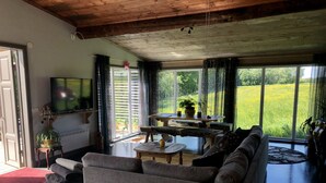 A living room with a sofa, a coffee table, and a TV. There is a window with curtains and a view of a green field. - Liljekonvalj Cottage Overlooking the River Sauna (Avesta)