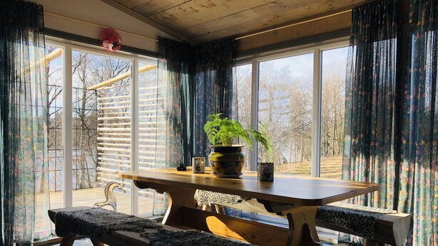 A room with a wooden table and benches, large windows with curtains, and a view of trees outside. - Liljekonvalj Cottage Overlooking the River Sauna (Avesta)