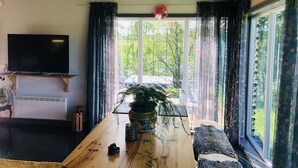 A living room with a wooden table, a flat-screen TV mounted on the wall, and a large window with curtains. - Liljekonvalj Cottage Overlooking the River Sauna (Avesta)