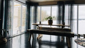 A room with a wooden ceiling, large windows with curtains, a wooden table with a plant, and a leopard-print bench. - Liljekonvalj Cottage Overlooking the River Sauna (Avesta)