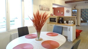 A dining area with a white table, red placemats, and a vase with red foliage. There is a kitchen area with a microwave, coffee maker, and toaster. The room has wooden beams on the ceiling and a window with a view outside. - Ravissant ! Le Panorama - Vue Mer (Criel-sur-Mer)