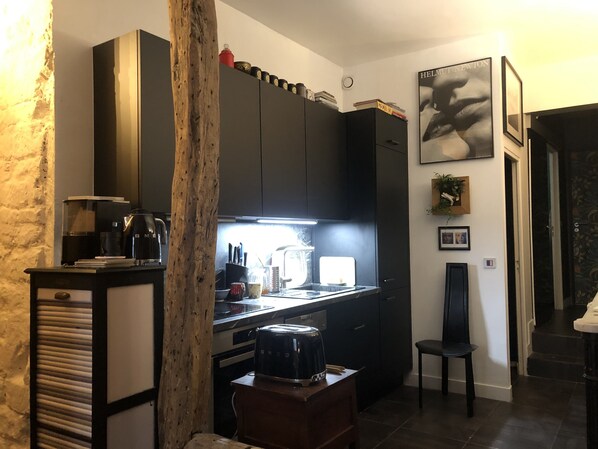Cozinha compacta com armários escuros, uma cafeteira e uma pequena área de refeições com uma cadeira.