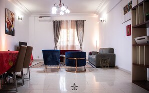 A modern living room with a dining area, featuring a chandelier, air conditioning unit, and a large window with patterned curtains. - Premier Residence at the heart of Dakar (Dakar)