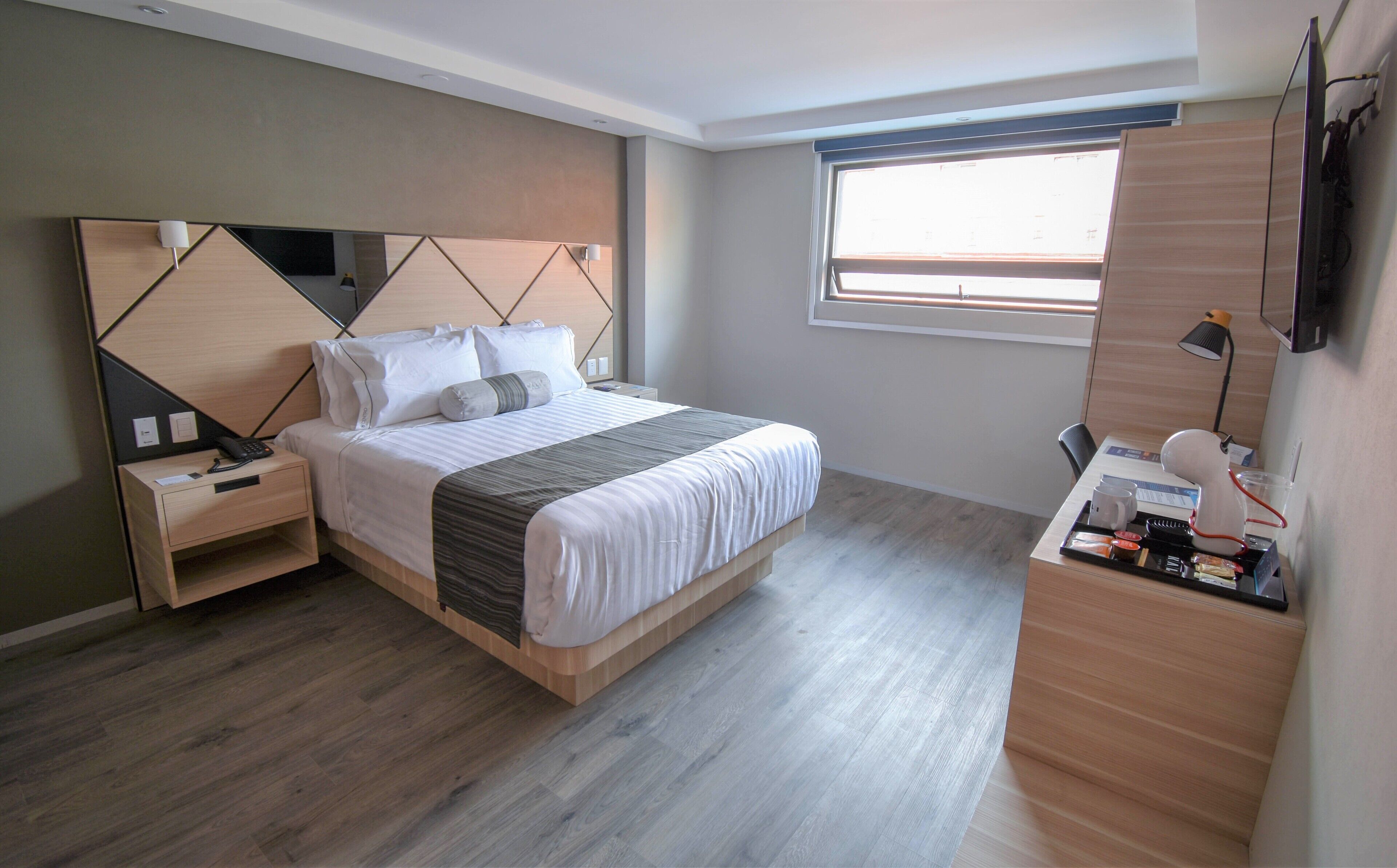 A modern bedroom with a wooden headboard, a bed with white bedding, a bedside table with a lamp, and a desk with a computer and a cup.