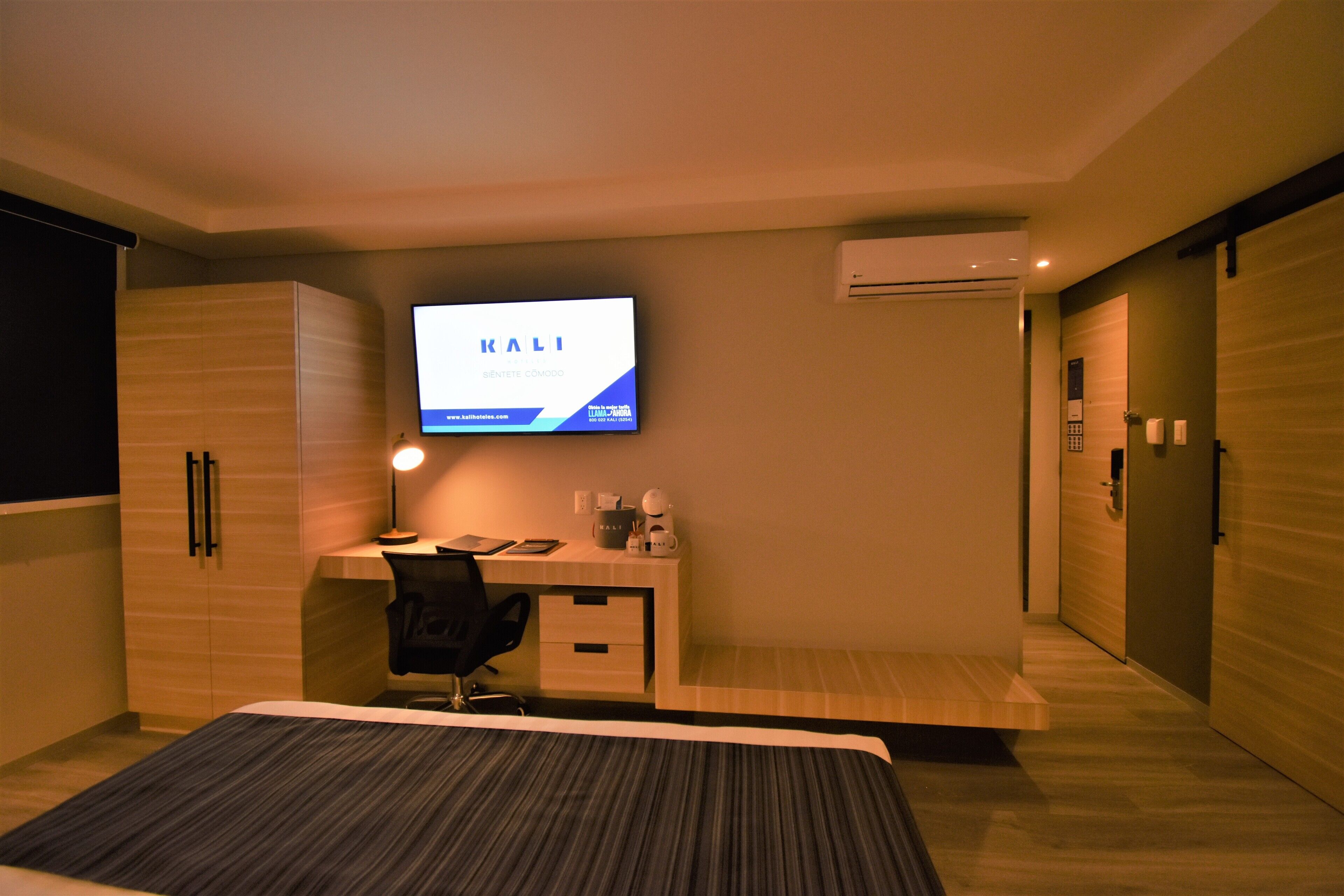 A modern hotel room with a bed, desk, and a TV mounted on the wall displaying 'KALI'.