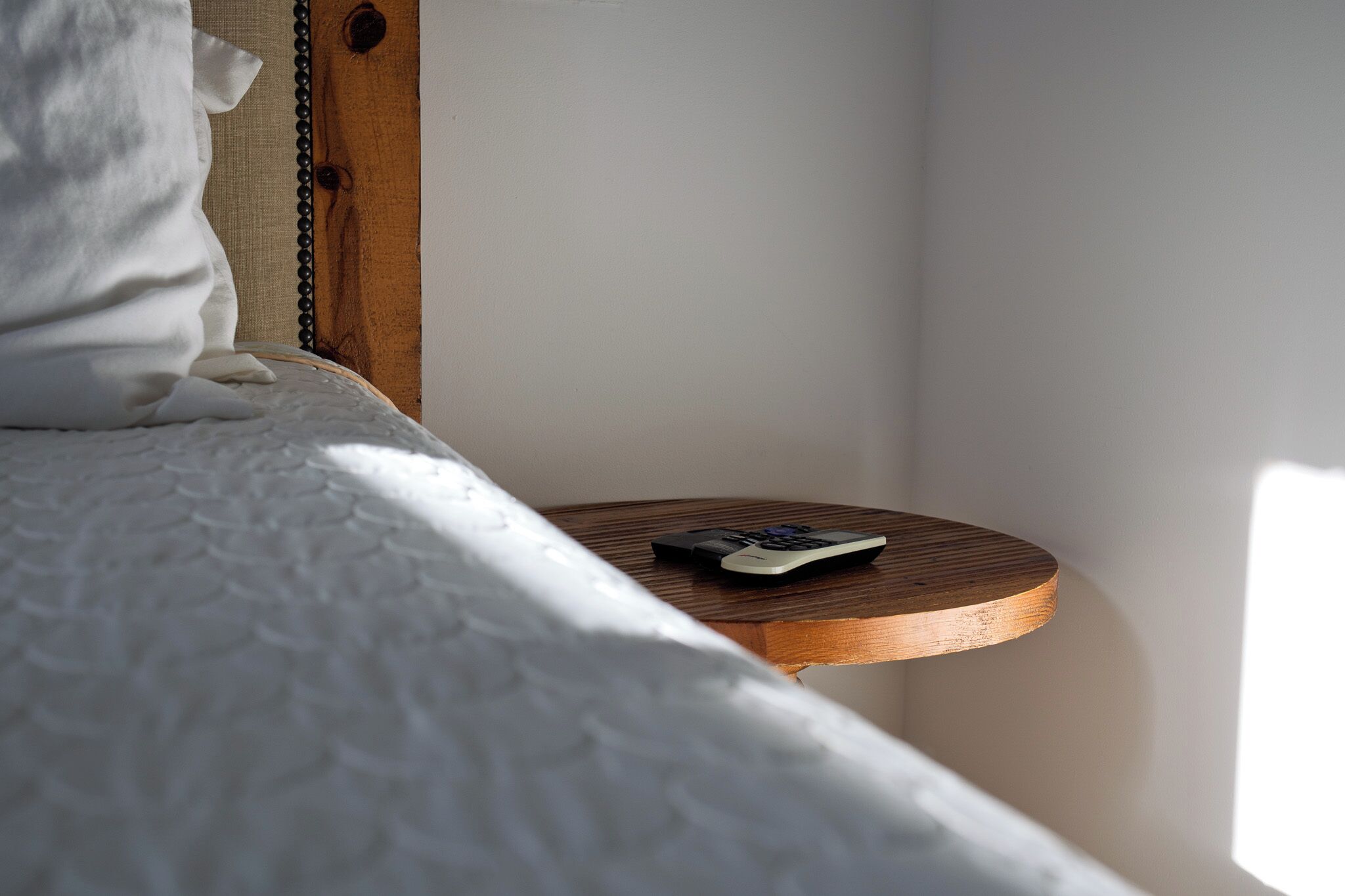 A bed with a white comforter and a remote control on a wooden bedside table.