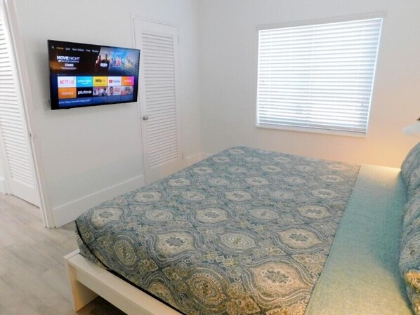 Ein Schlafzimmer mit einem Bett, einem an der Wand montierten Fernseher, einem Fenster mit Jalousien und einer Tür.