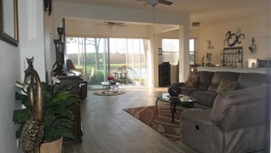A spacious living room with a leather sofa, a coffee table, and a dining area with chairs. There is a large window providing natural light and a view of the outdoors. - Relax on your lanai after a round of golf and enjoy our beautiful SWFL sunsets! (Fort Myers)